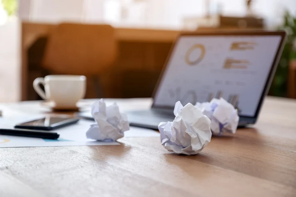 Crumpled paper balls on a desk beside a laptop and coffee cup representing productivity frustration
