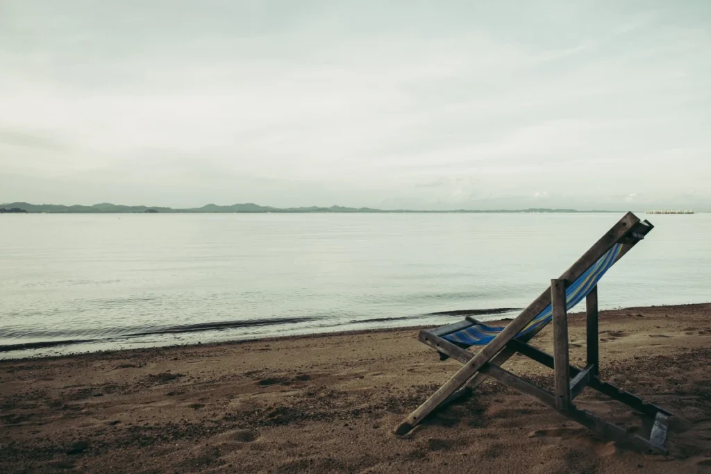 Lone deck chair on a quiet beach under an overcast sky representing the question of recharging versus collapsing on vacation