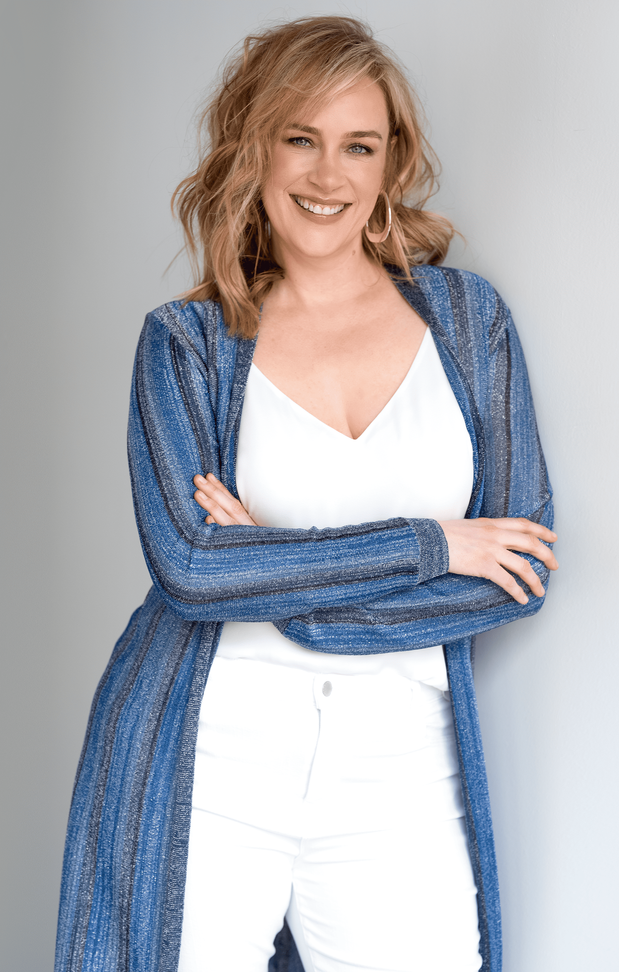 a picture of allison graham leaning against a grey wall wearing a blue striped cardigan, white jeans and a white top. she is smiling and her hair is wavy