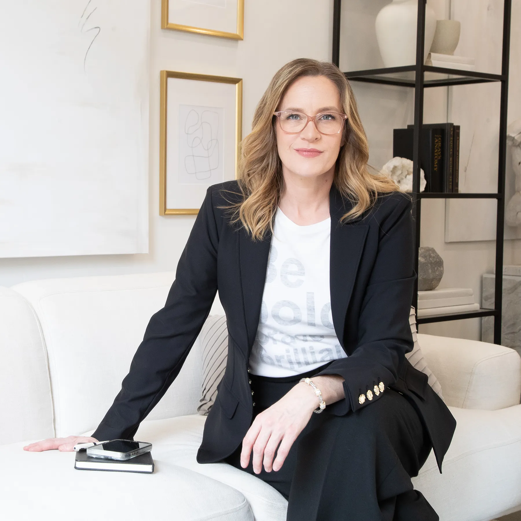 Allison Graham sitting on a cream coloured couch in a black suit and a white tshirt that says be bold be brave be brilliant